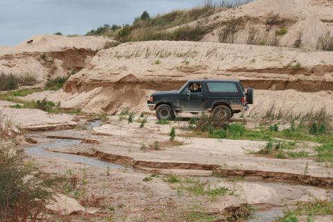 1987 Ford Bronco - A 1987 Ford Bronco navigates a dry, sandy wash with eroded banks under a cloudy sky.