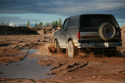 1987 Ford Bronco - A black 1987 Ford Bronco kicks up mud as it drives through a watery track in a sandy area.