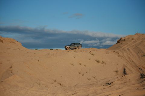 1987 Ford Bronco - A 1987 Ford Bronco drives across a sandy landscape with a dramatic sky overhead.