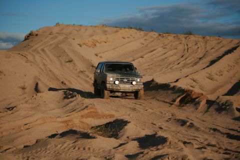 1987 Ford Bronco - A 4x4 vehicle navigates a steep sand dune under a partly cloudy sky.