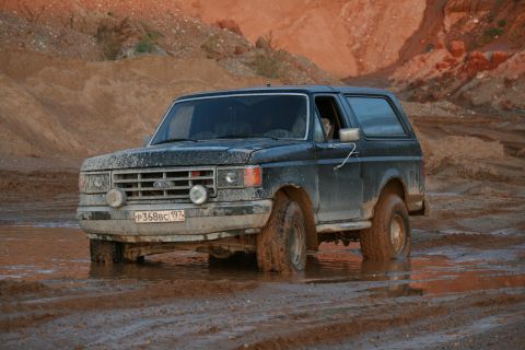1987 Ford Bronco - A dark blue 1987 Ford Bronco navigates a muddy terrain, with its front-left tire deep in the mire.