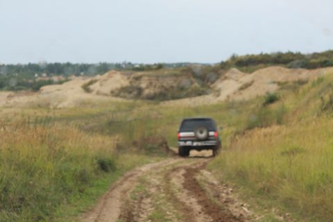 1987 Ford Bronco - SUV navigates a dirt track through a grassy, hilly landscape. SUPERMOTORS.NET logo visible.