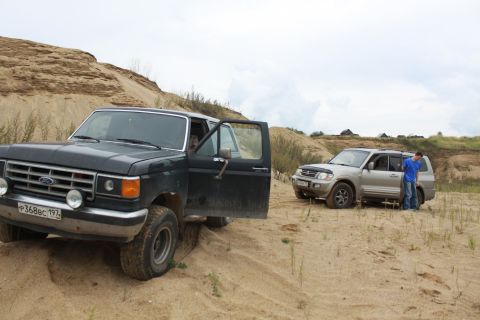 1987 Ford Bronco - A dark green 1987 Ford Bronco is parked on a sandy hill with its driver's door open, while another SUV is visible in the background.