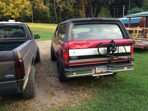 1994 Ford Bronco - Two Ford trucks are parked side-by-side on a gravel and grass area, with trees and a building in the background.