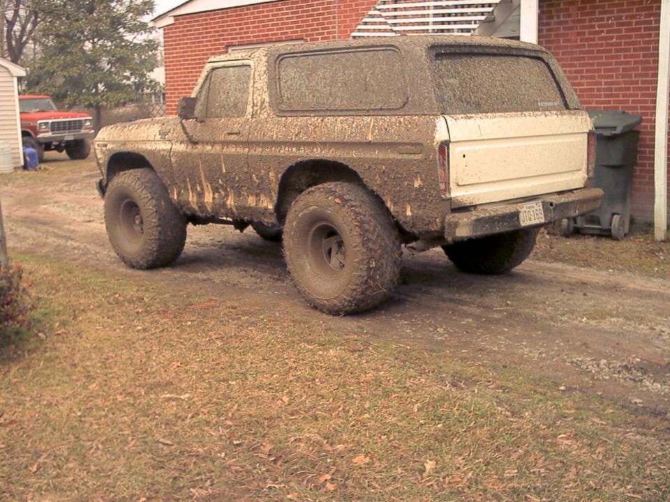 Mud-covered lifted Ford Bronco parked on grass beside a brick house, rear three-quarter view