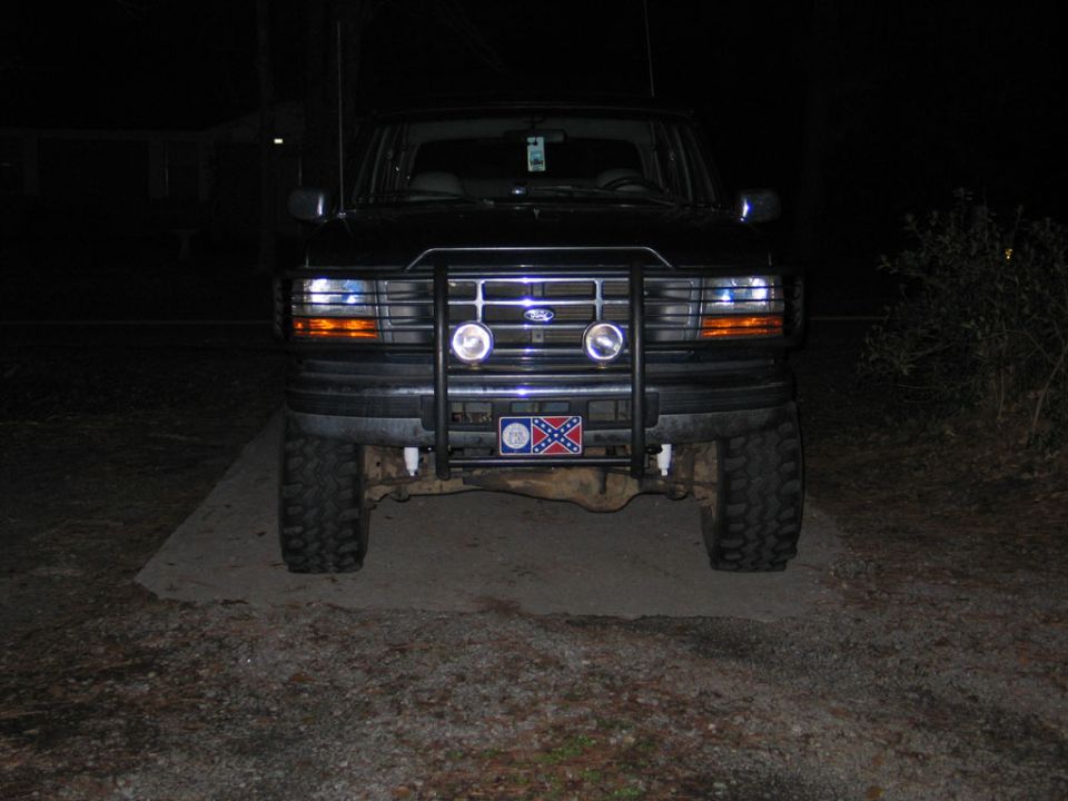 Front view at night of lifted dark Ford Bronco with grille guard, driving lights, mud tires