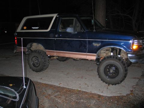 1995 Ford Bronco - A lifted blue Ford Bronco sits on a paved surface at night, showing off its off-road tires and suspension.