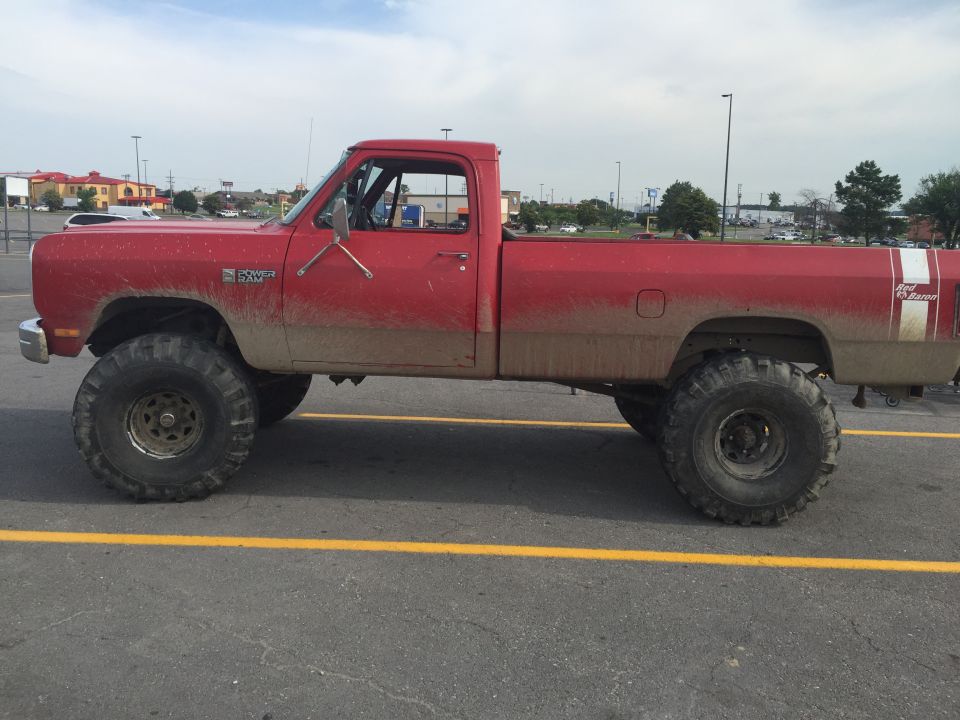 Red 1987 Dodge Power Ram pickup truck with large off-road tires, mud splattered on the lower body, parked on asphalt