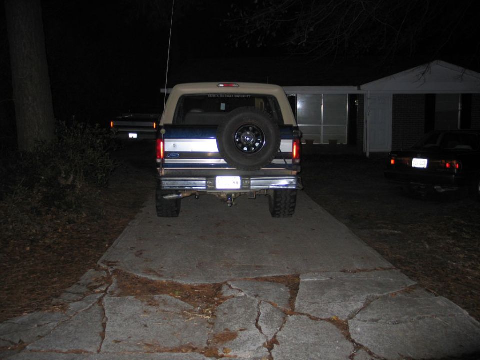 Rear view at night of a lifted Ford Bronco with tan top and tailgate-mounted spare tire