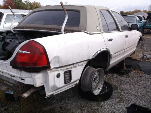 1999 Mercury Grand Marquis - Rear view of a white 1999 Mercury Grand Marquis in a junkyard, missing a rear wheel and with a damaged bumper.