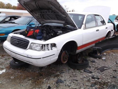 1999 Mercury Grand Marquis - White sedan with its hood open, missing wheels and front bumper, sits in a junkyard.