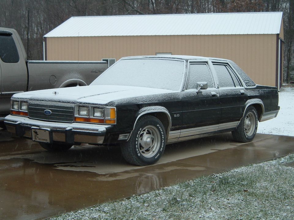 Black Ford LTD Crown Victoria sedan with light snow in a driveway by a barn