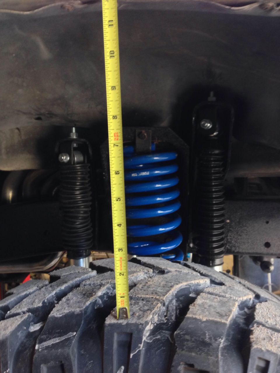 Close-up of a blue coil spring and black shock absorber with a yellow measuring tape next to a large tire