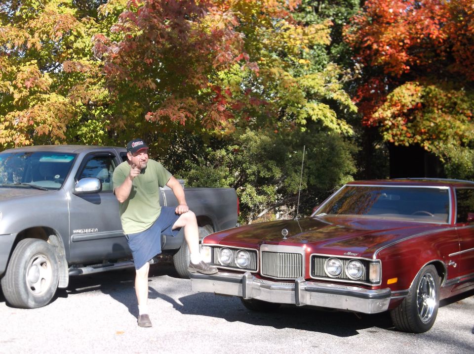 Man standing between a silver pickup truck and a maroon 1977 Mercury Cougar XR-7