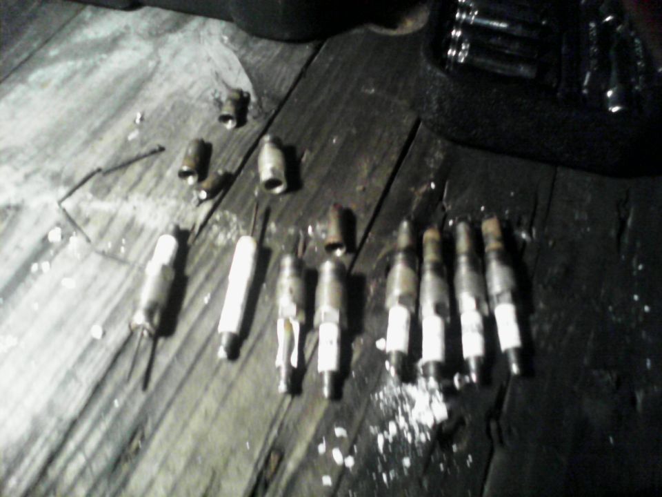 Eight used spark plugs and loose sockets laid out on a dark wooden floor beside a socket set.