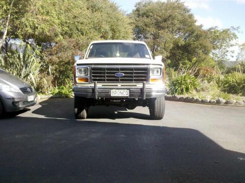 1982 Ford Bronco - A white Ford Bronco is parked in front of trees and foliage, with another vehicle to its left.