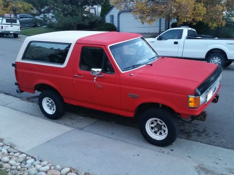 1988 Ford Bronco - A red Ford Bronco with a white top is parked on a street, with a white pickup truck behind it.