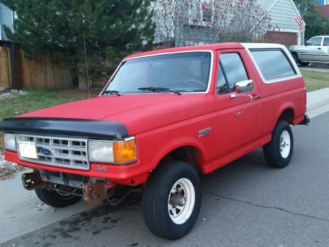 1988 Ford Bronco - This red Ford Bronco is parked on a residential street, showing its front three-quarter view with a white top.
