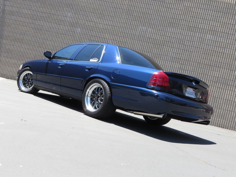 Dark blue lowered Ford Crown Victoria sedan with chrome wheels by a corrugated metal wall
