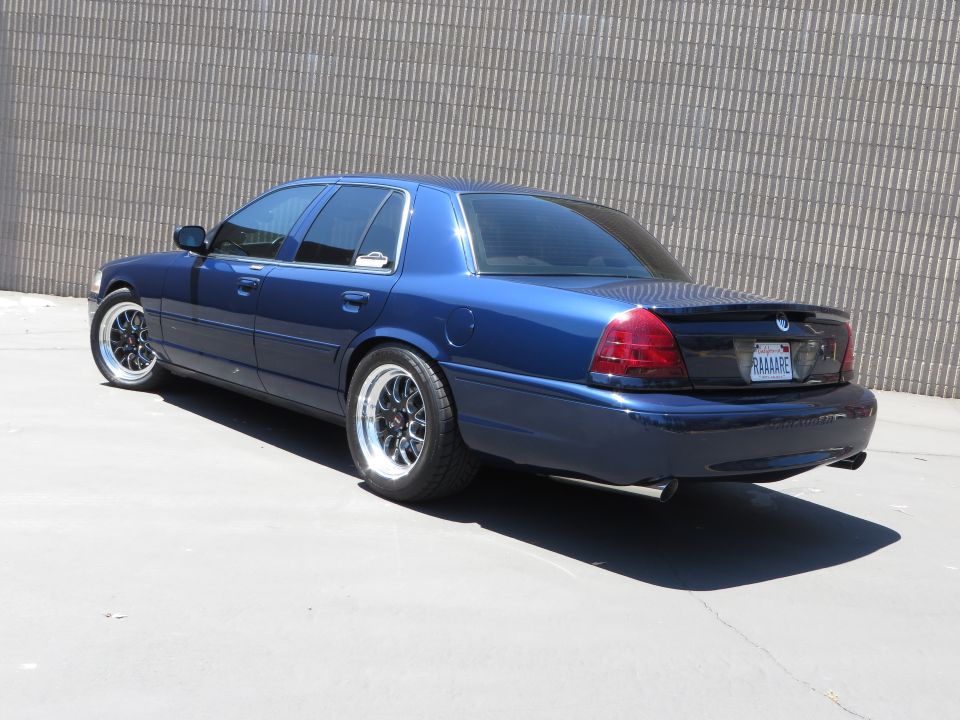 Dark blue Mercury Grand Marquis sedan with chrome wire wheels parked beside a metal wall