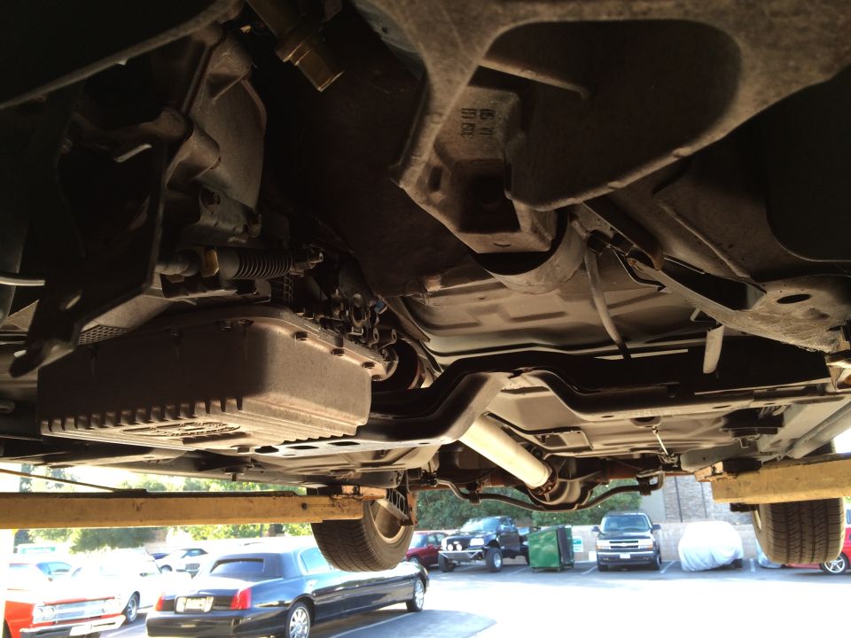 Underside view of a car on a lift, showing the transmission and exhaust. Other cars are parked in the background