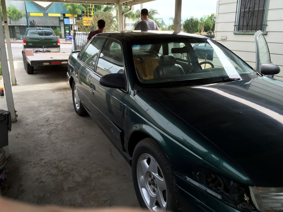 Dark green sedan parked under a carport, with a green pickup truck and trailer behind it