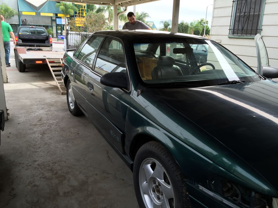 Dark green sedan parked under a carport, with a pickup truck and trailer in the background