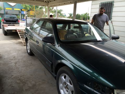 1993 Ford Taurus - A green sedan is parked under a carport, with a man opening the driver's door as a black pickup truck is loaded onto a trailer nearby.