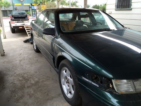 1993 Ford Taurus - 1993 Ford Taurus parked next to a trailer with a pickup truck. The Taurus is missing its driver's side headlight.