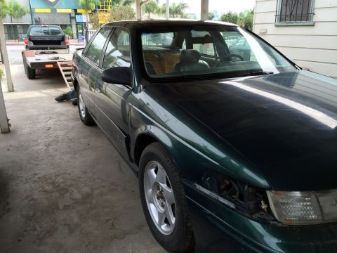 1993 Ford Taurus - A dark green sedan is parked outdoors, with a black pickup truck on a trailer in the background.