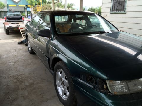 1993 Ford Taurus - Dark green 1993 Ford Taurus sedan with a missing headlight, parked next to a trailer with a pickup truck.