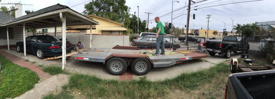 Dark sedan being loaded onto a trailer under a carport, with a black pickup truck nearby