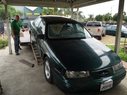1993 Ford Taurus - A man is loading a green sedan onto a trailer. A pickup truck and another car are parked nearby.