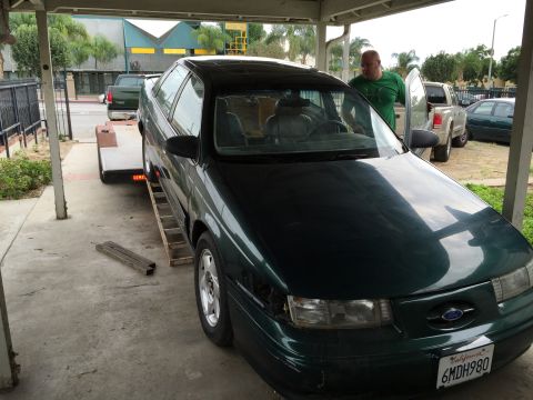 1993 Ford Taurus - A man is loading a dark green sedan onto a trailer, with other vehicles visible in the background.