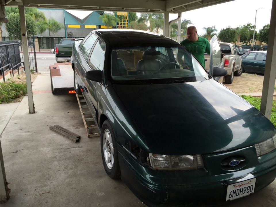 Dark green sedan being loaded onto a trailer. A man in a green shirt stands by the open car door