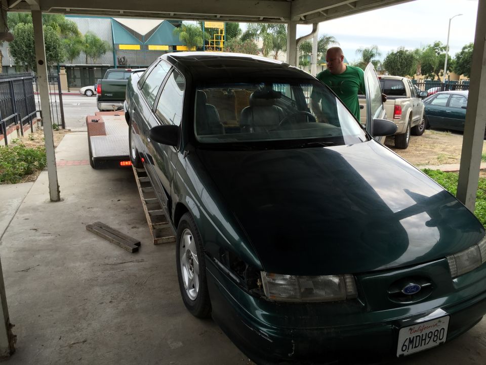 Dark green sedan on a trailer, front right corner damaged, man standing by open door