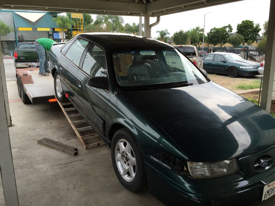 Dark green sedan being loaded onto a trailer. Another sedan is parked in the background