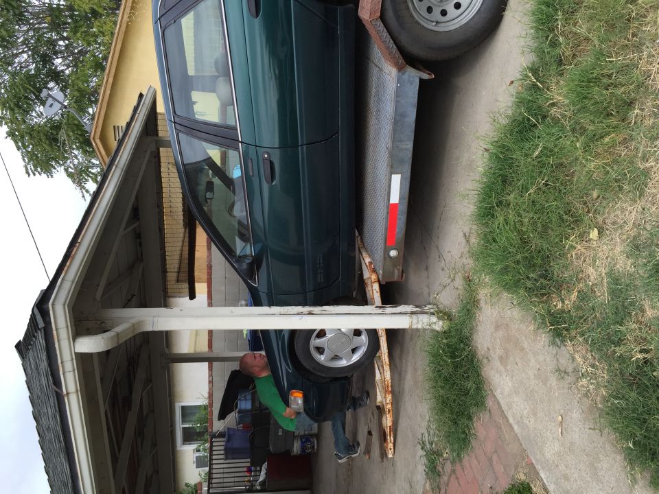 Green sedan on a trailer, being loaded under a carport. Side view
