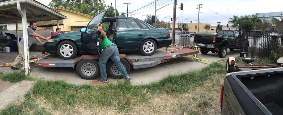 Green sedan on a trailer being pushed by two men, with a black pickup truck in the background