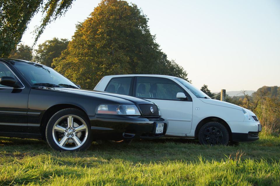 Black Mercury Grand Marquis parked next to a white Volkswagen Lupo on a grassy field