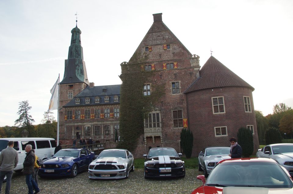 Several Ford Mustangs are parked in a lot in front of a large brick castle with a tall tower