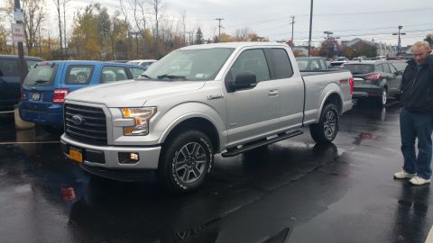 2015 Ford F-150 - A silver Ford F-150 is parked on a wet surface, with other vehicles and a man in the background.