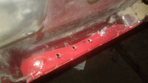1996 Ford Bronco - Detail of a red painted metal part showing multiple drilled holes and surface imperfections.