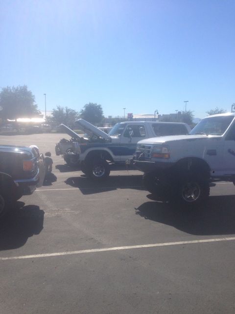 1978 Ford Bronco - Several lifted SUVs are parked in a lot under a clear blue sky, with one vehicle's hood open.