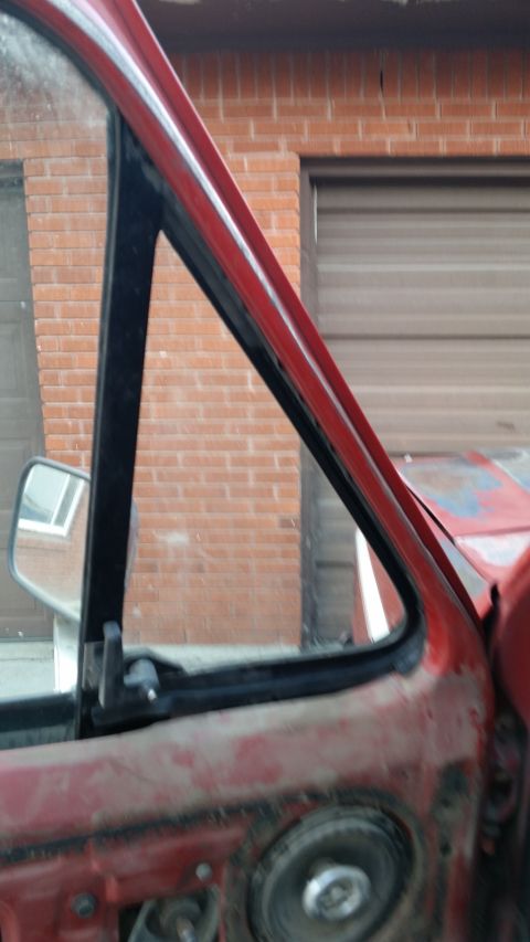 1981 Ford Bronco - Close-up view of a red truck's door frame and side mirror, with a brick building and garage door visible behind it.