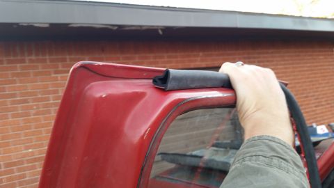 1981 Ford Bronco - A hand holds a piece of weatherstripping along the top edge of a red vehicle's window frame.