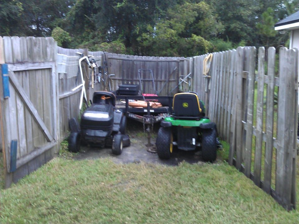 Two riding lawn mowers, one black and one green John Deere, parked next to a utility trailer in a fenced yard