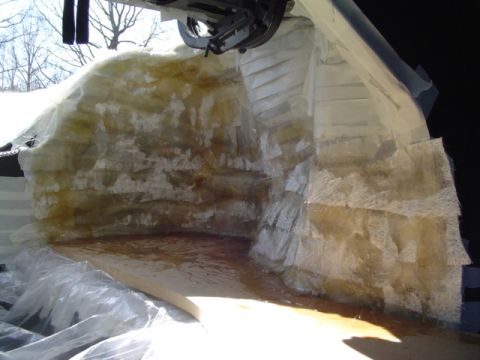 1999 Ford Taurus - Fiberglass mat being laid up over the trunk's rear bulkhead and quarter panels, with wet resin pooling amber on the MDF floor below and cle…