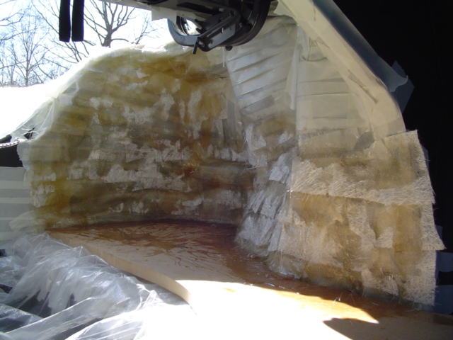 Fiberglass mat laid up over a trunk bulkhead with wet resin pooling on the MDF floor