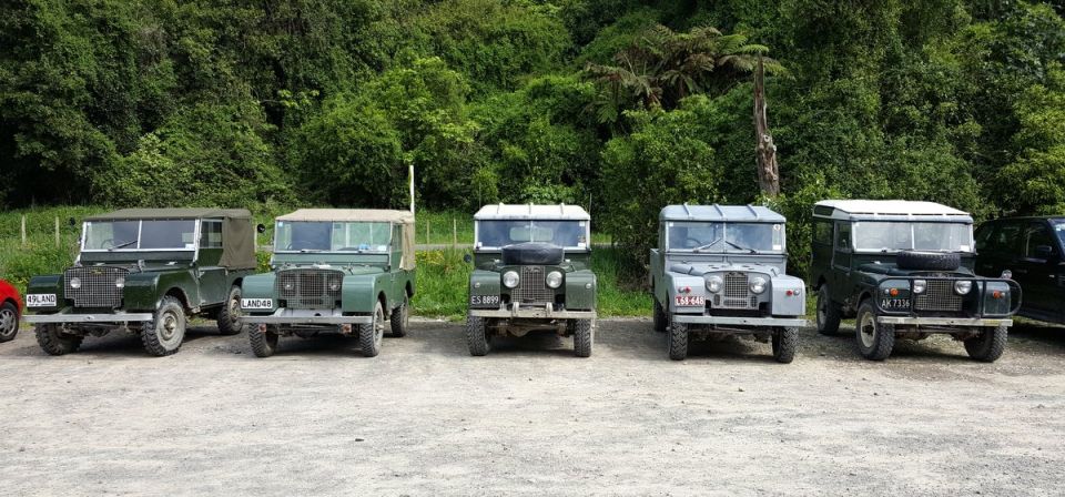 Five vintage Land Rovers parked outdoors with trees in the background. License plates are visible